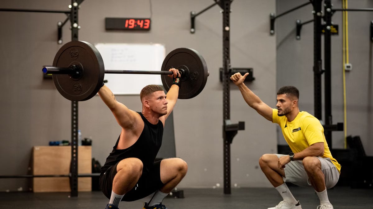 Athlete performing weightlifting exercise under trainer's guidance in a gym setting.